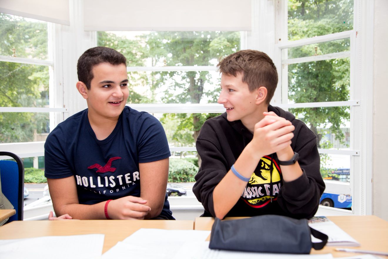 Two teenage students working together at a desk beside a classroom window during an English lesson