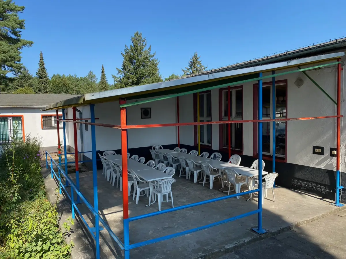 A single-storey camp building with a covered terrace and stacked white chairs along the front