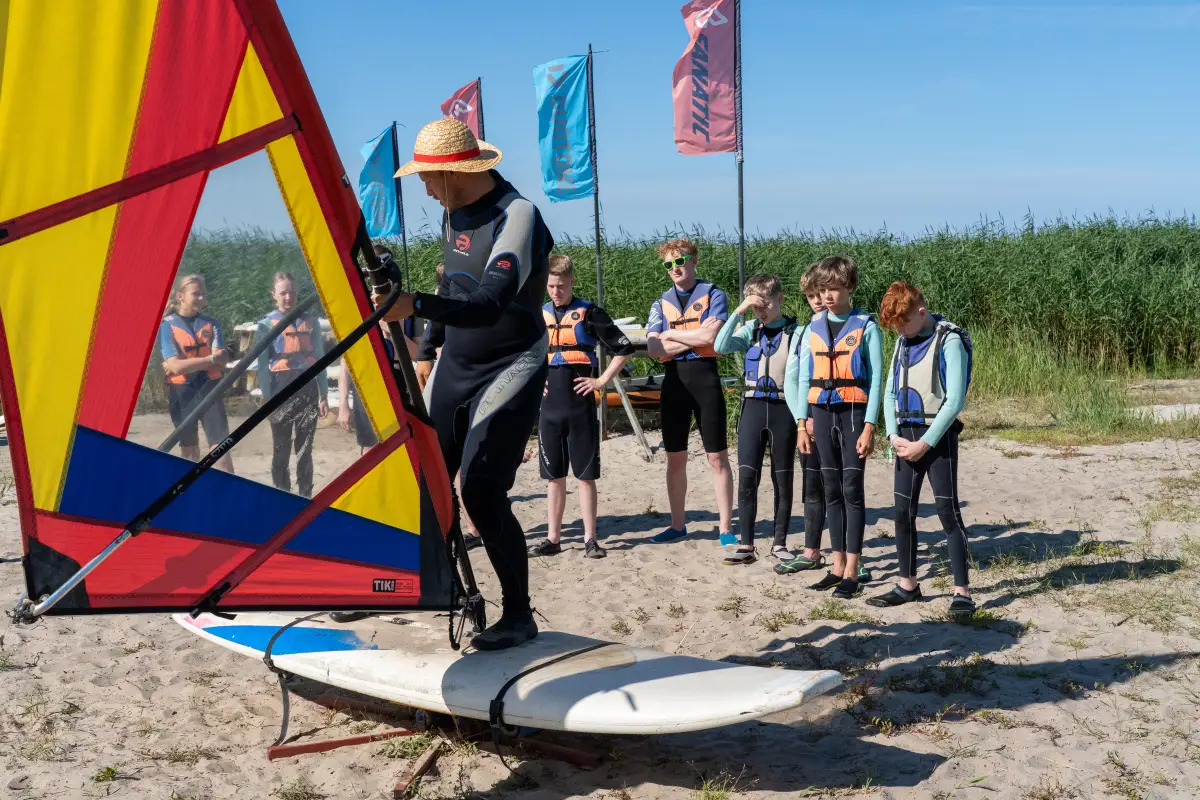 A group gathered on the sand beside a rigged windsurf board, flags along the beach behind them
