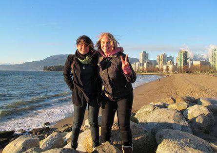 Two students standing on a pebble beach with the Vancouver skyline and the bay behind them
