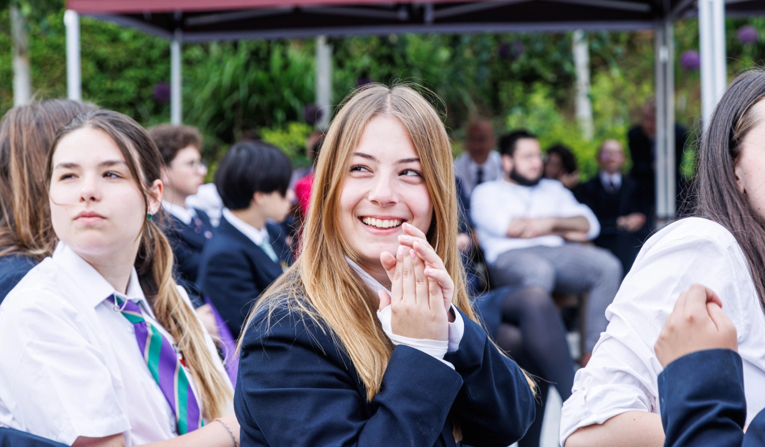 Students in school uniform seated outdoors under a canopy at a campus event, Mill Hill School, London