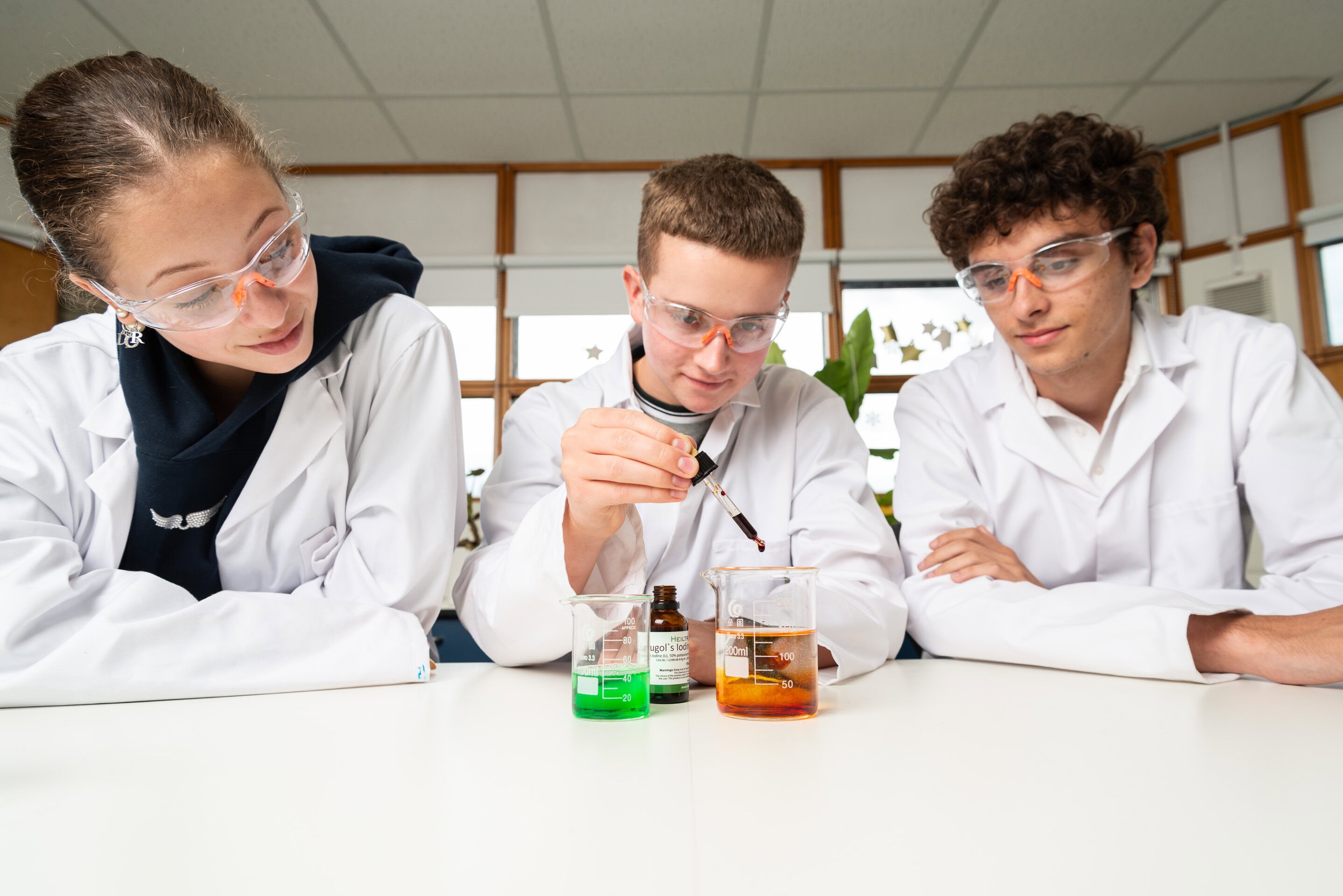 Three students in lab coats and safety goggles work with coloured liquids during a science session