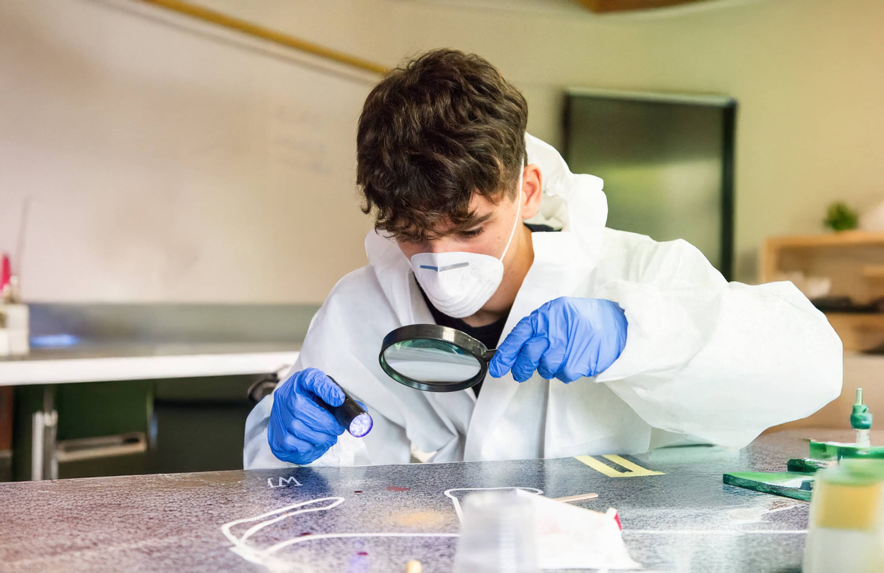 A student in a lab coat, mask and gloves examines a sample with a magnifying glass at a laboratory bench