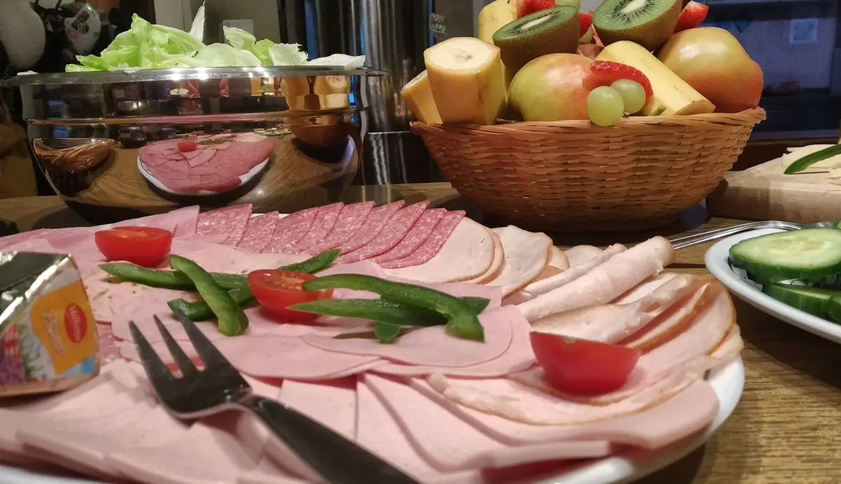 A buffet platter of sliced meats and cheese beside baskets of apples and kiwi fruit