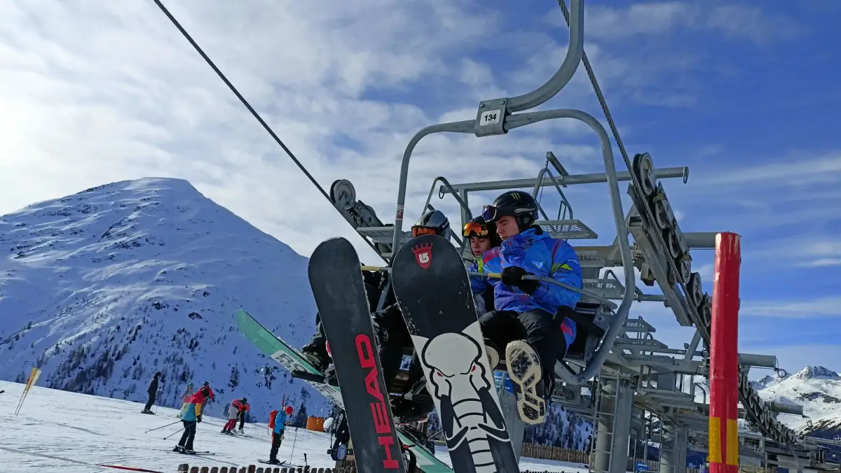 Two participants with skis and a snowboard on a chairlift, with a snow-covered ridge below