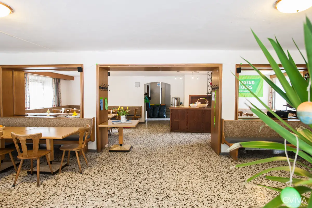 A plain dining room with wooden tables and chairs opening through to a serving area