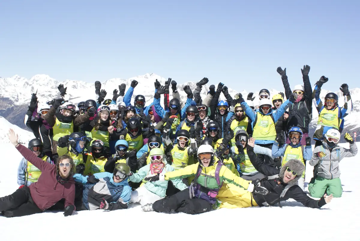 A large group of young skiers and snowboarders in ski gear posing together on a sunlit slope with mountains behind