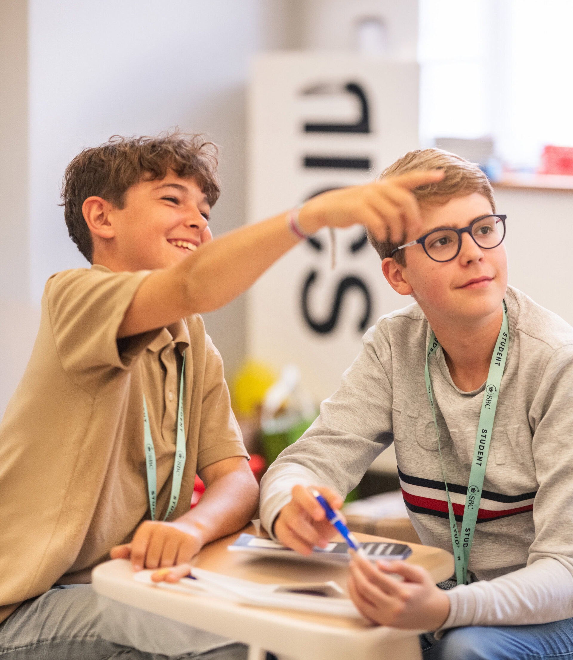 A student points ahead while another sits beside him with a pen at a table