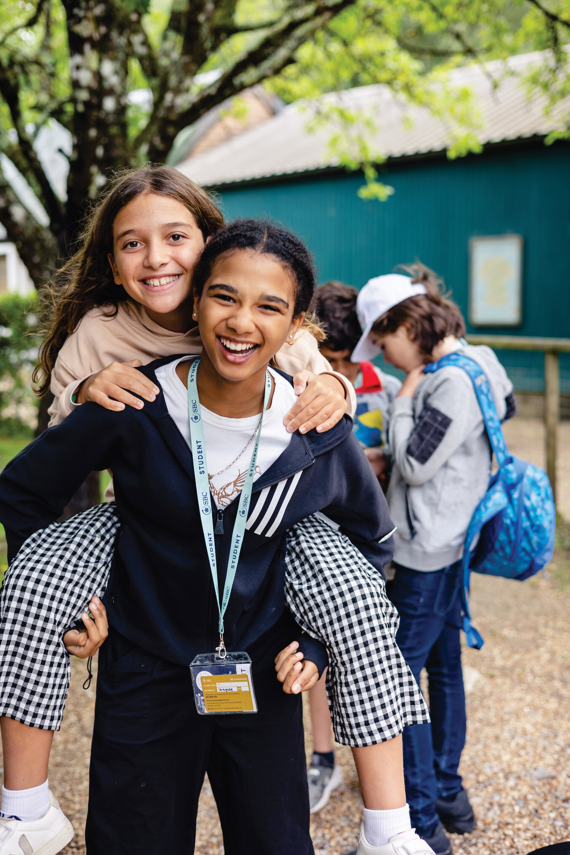 A student gives another a piggyback ride outside, both smiling and wearing camp clothing