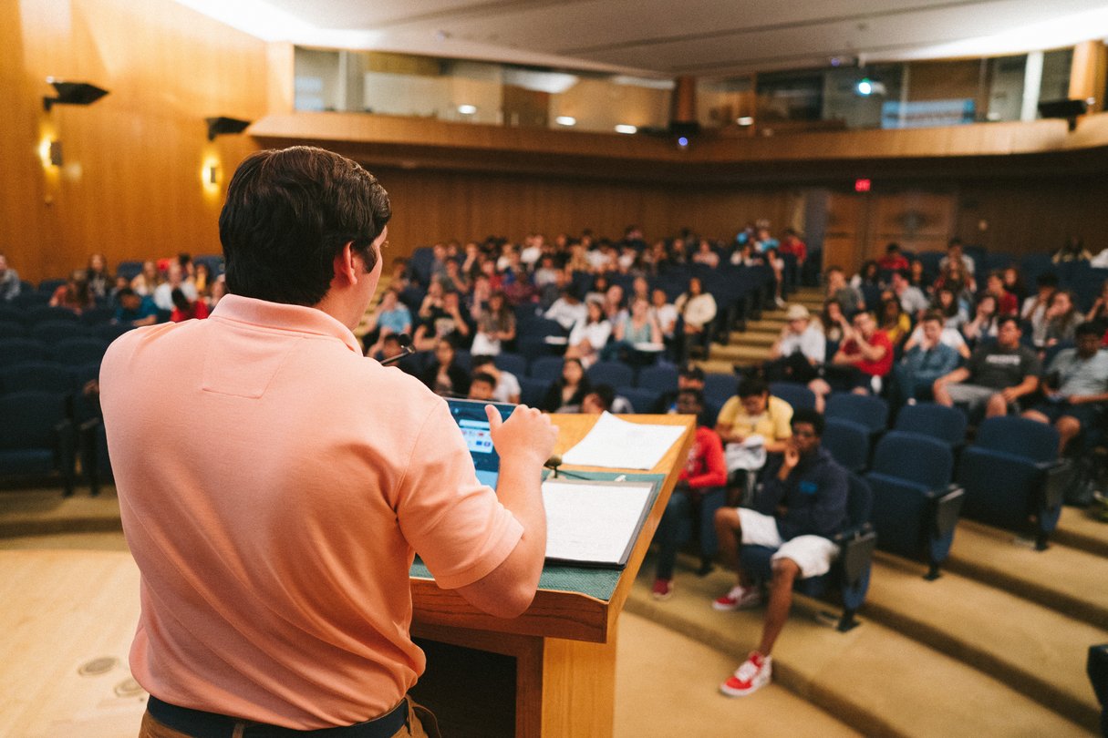 A lecturer speaking from the podium to a full tiered lecture hall of summer programme students