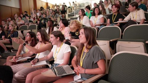 Students in a tiered lecture hall watching a presentation, notebooks and laptops open