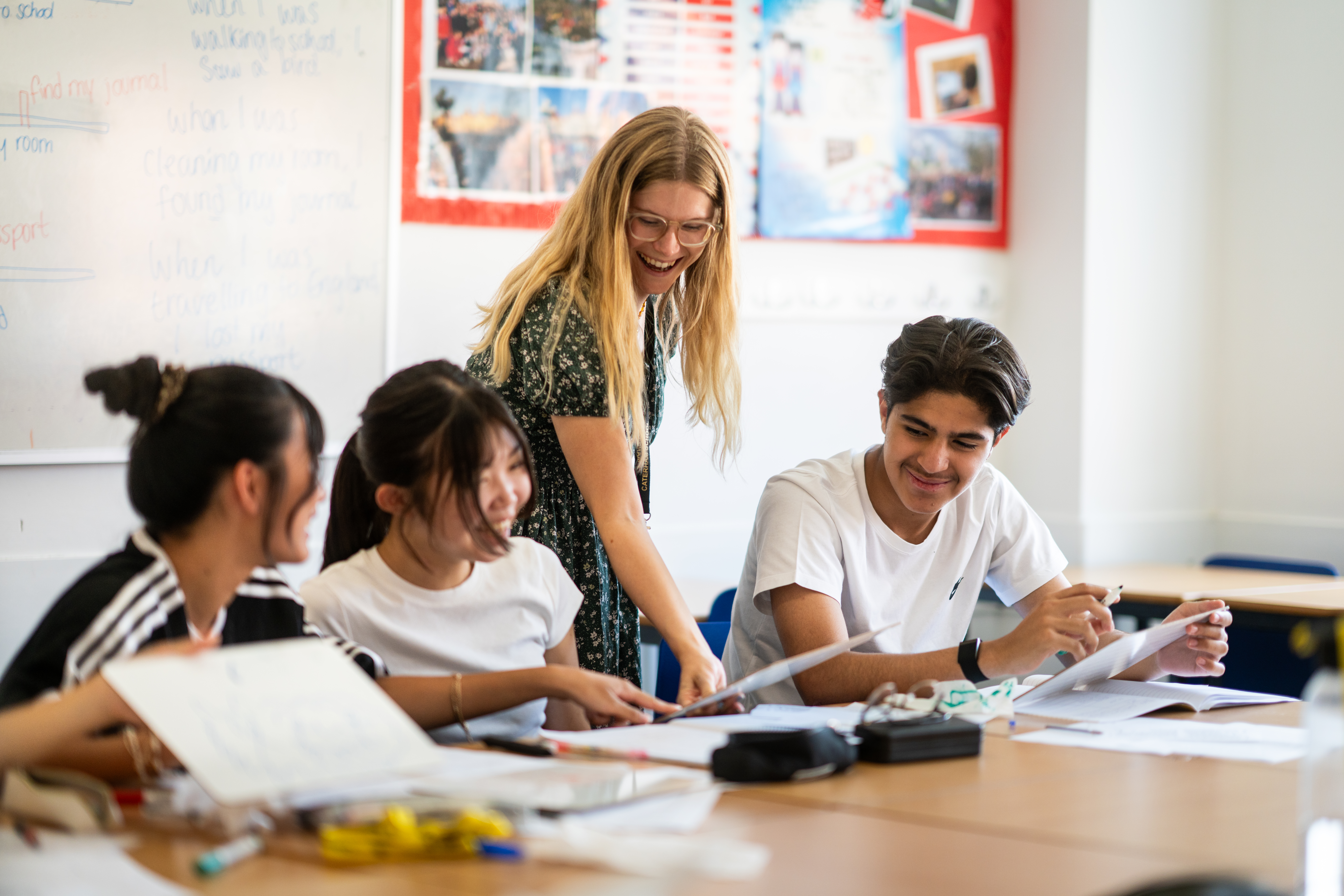Teacher leaning over a table of four students working through printed worksheets in a bright classroom