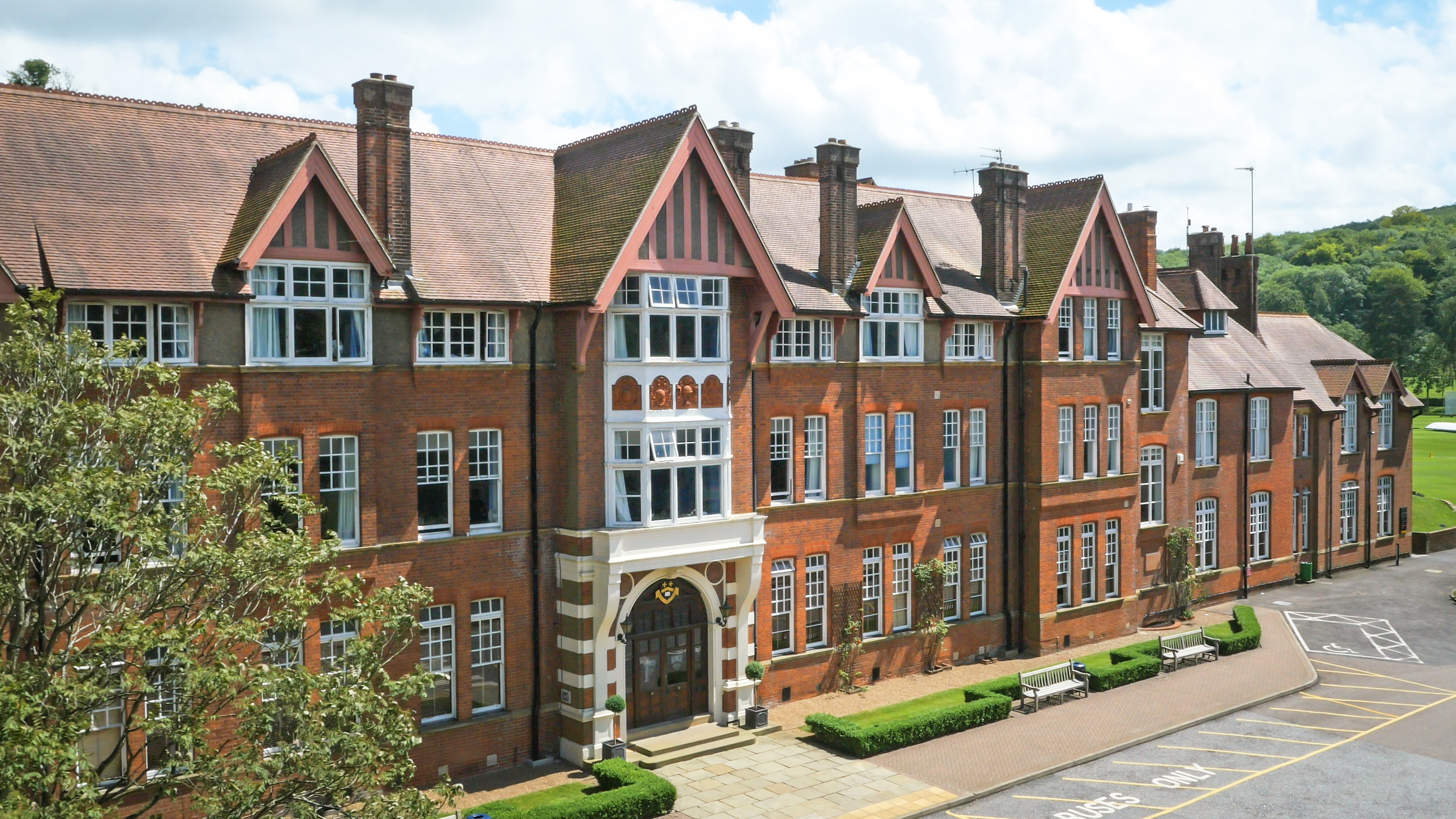 Red-brick Victorian school building with tall gables and bay windows, lawn and mature trees in front
