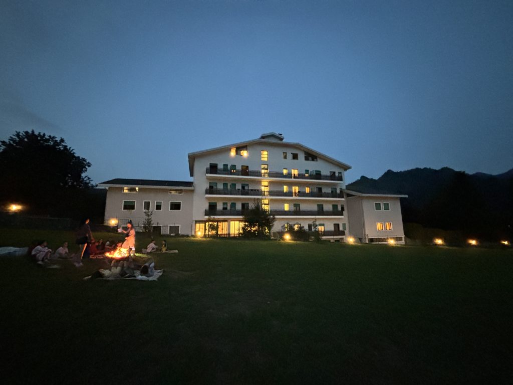 Villa Sancelso lit up at dusk, seen across its lawn, with children sitting on the grass around a fire in the foreground and wooded hills behind