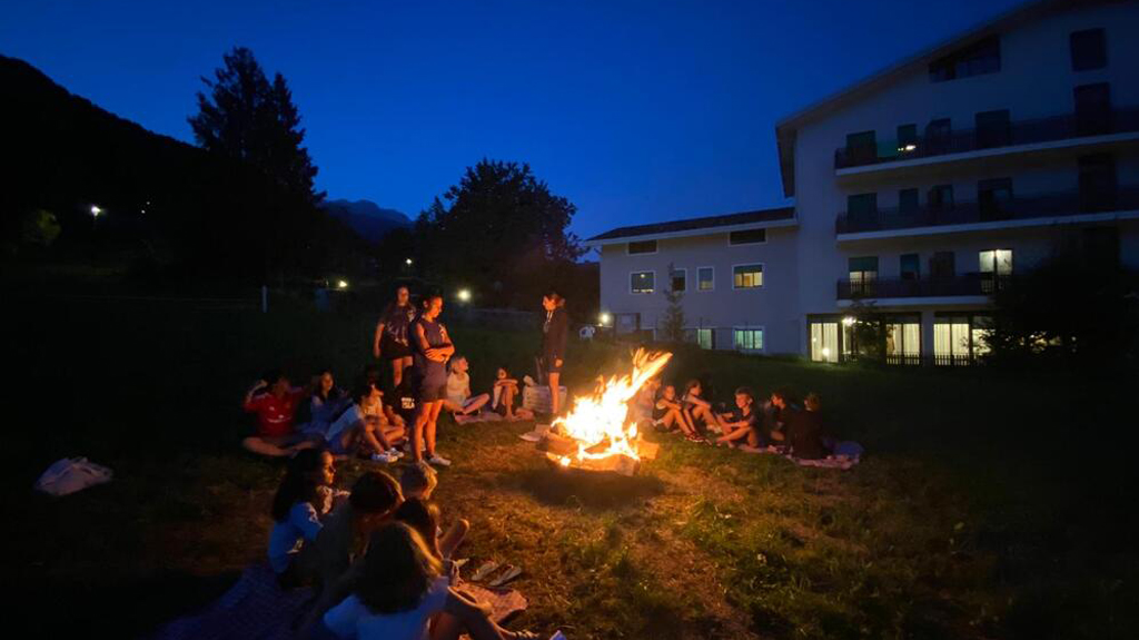 Children and staff sitting in a wide circle on the grass around a bonfire at night, with the lit windows of Villa Sancelso behind them