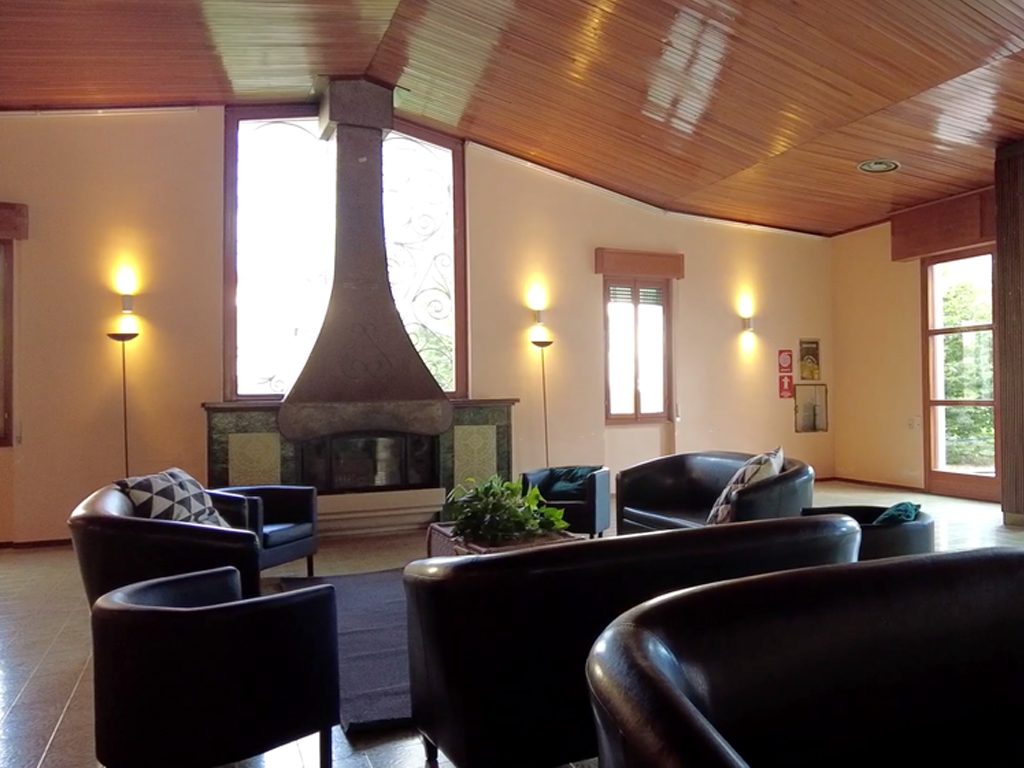 A common room at Villa Sancelso with dark sofas and armchairs grouped around a tall conical fireplace, under a wood-panelled ceiling