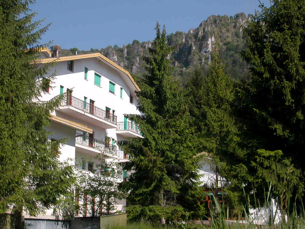 The white facade of Villa Sancelso with green shutters and balconies, framed by conifers, with the rock of the Presolana rising behind it