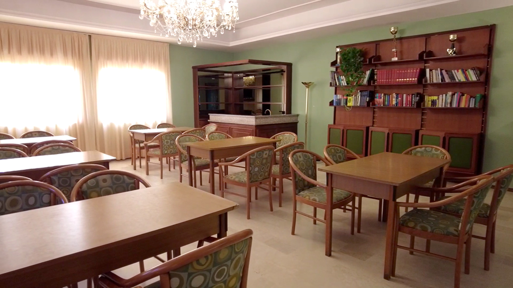 A long room at Villa Sancelso set with wooden tables and upholstered chairs, bookshelves along the far wall and tall curtained windows letting in daylight