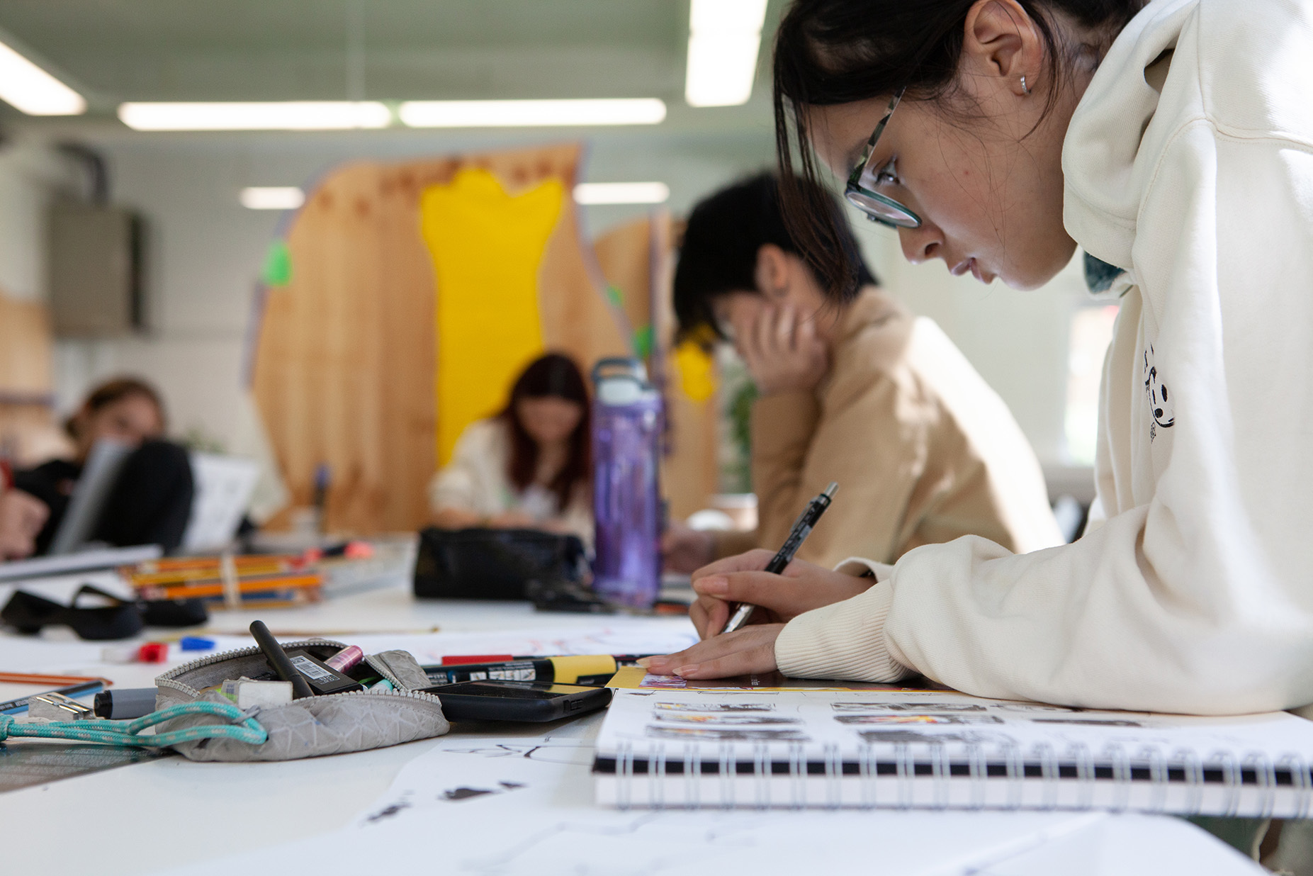 Participants sitting at studio tables drawing in sketchbooks, with pens and markers laid out in front of them