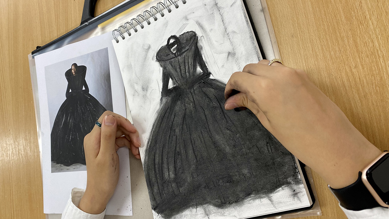 Hands working on a charcoal drawing of a black dress in a sketchbook beside a printed reference photograph