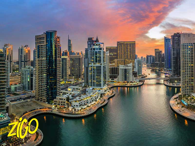 High-rise towers lining the Dubai Marina waterway at sunset, with boats moored along the channel