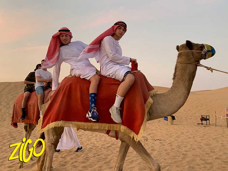 Participants in white robes and headscarves riding a camel led across the sand